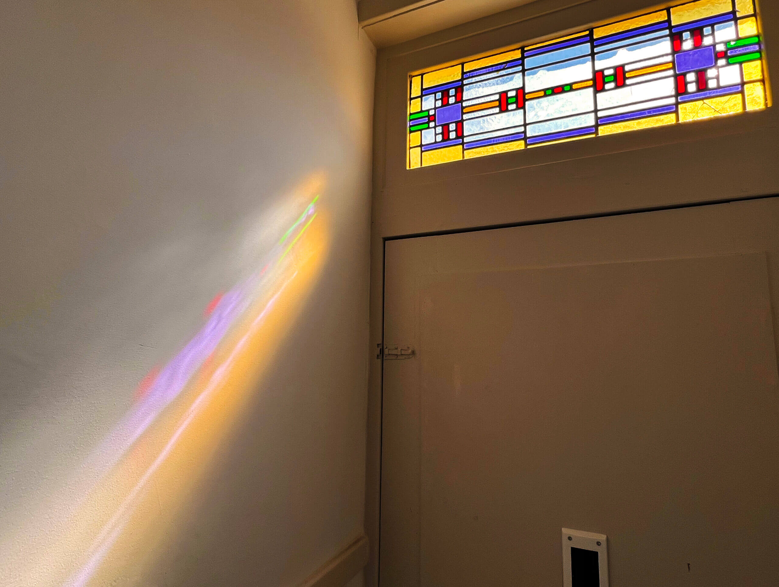 A stained-glass window above a closed doorway with sunlight streaming in